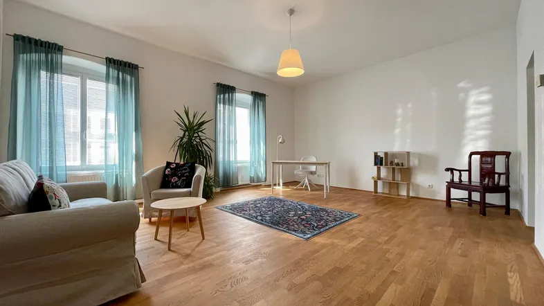 Geräumiges Wohnzimmer mit Holzboden, zwei Fenstern und einem Arbeitsbereich im Hintergrund.