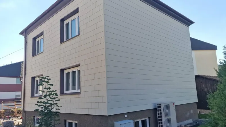 Modernes Gebäude mit heller Fassadenverkleidung und sichtbarer Wärmepumpe im Außenbereich.