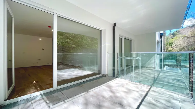 Heller Balkon mit Fliesenboden und Glasgeländer, bietet Zugang zu einem leeren Raum.