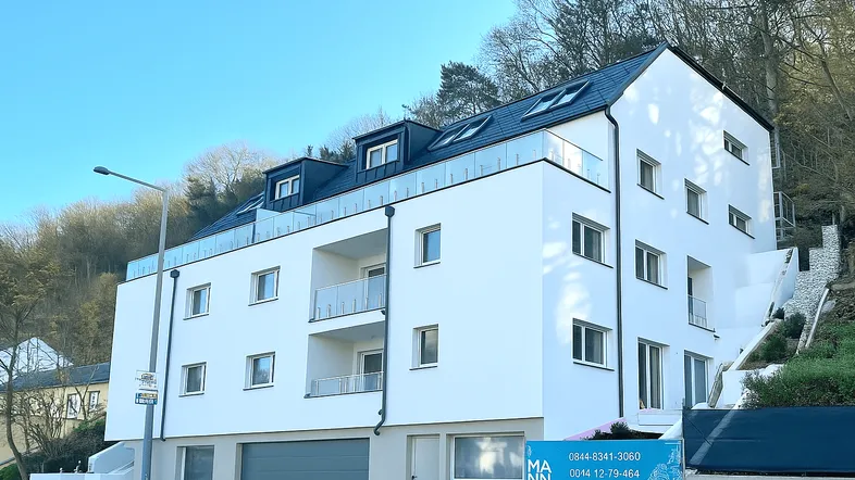 Modernes Mehrfamilienhaus mit weißer Fassade, Balkonen und Dachgauben, eingebettet in eine grüne Hanglage.