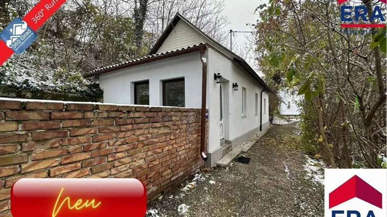 Weißes Einfamilienhaus mit Satteldach und Ziegelmauer im Vordergrund, umgeben von Bäumen.