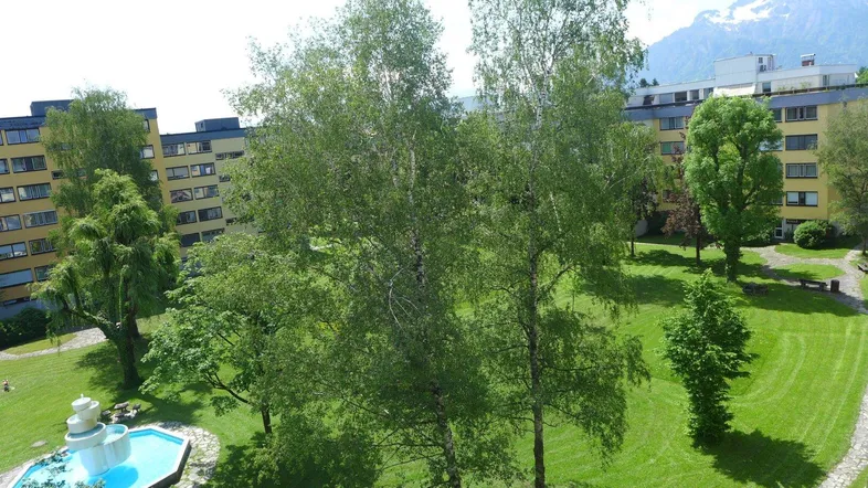 Grüner Innenhof mit Springbrunnen, umgeben von Wohngebäuden und mit Bergblick.