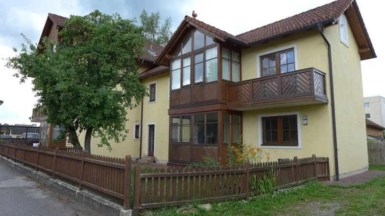 Gelbes Mehrfamilienhaus mit Holzzaun und Balkonen in ländlicher Umgebung unter bewölktem Himmel.