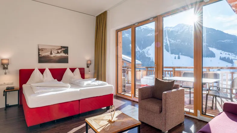 Helles Schlafzimmer mit rotem Bett, Holzboden und großem Balkonzugang mit Bergblick.