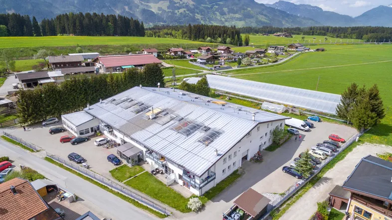 Luftaufnahme eines großen Gebäudekomplexes mit Parkplätzen und umliegenden Feldern vor Bergkulisse.