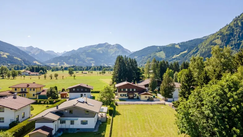 Luftaufnahme einer Wohnsiedlung mit Häusern, grünen Wiesen und majestätischen Bergen im Hintergrund unter blauem Himmel.