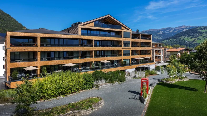 Modernes Gebäude mit Holzfassade, vielen Balkonen und gepflegter Gartenanlage vor alpiner Bergkulisse.
