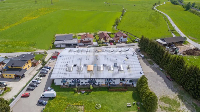 Luftaufnahme eines Gebäudekomplexes mit großem Gartenbereich, Parkplätzen und umliegender grüner Landschaft.
