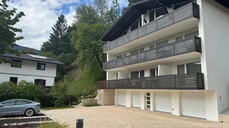 Modernes Mehrfamilienhaus mit mehreren Balkonen und Garagen, umgeben von grüner Landschaft und Bergen.