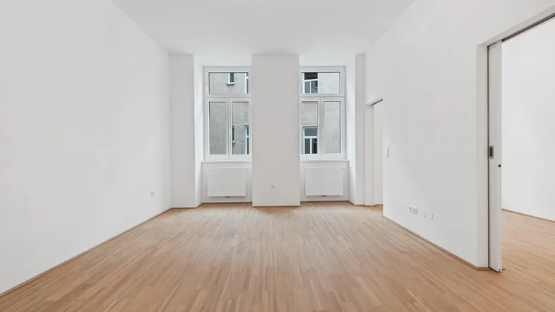 Helles, leeres Zimmer mit Parkettboden und zwei Fenstern, bereit zur individuellen Gestaltung.