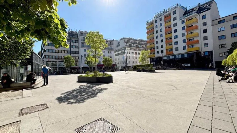 Ein sonniger Stadtplatz mit modernen Gebäuden, Bäumen und Sitzgelegenheiten, der eine lebendige Umgebung zeigt.