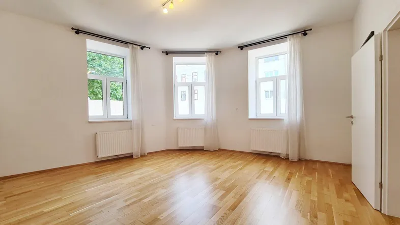 Helles Zimmer mit Parkettboden, weißen Wänden und drei großen Fenstern, die viel Licht hereinlassen.