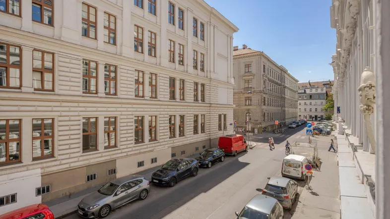 Blick auf eine belebte Stadtstraße mit geparkten Autos und historischen Gebäuden unter blauem Himmel.