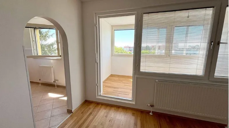 Helles Zimmer mit Parkettboden, Zugang zu einem verglasten Balkon und einem angrenzenden Raum.