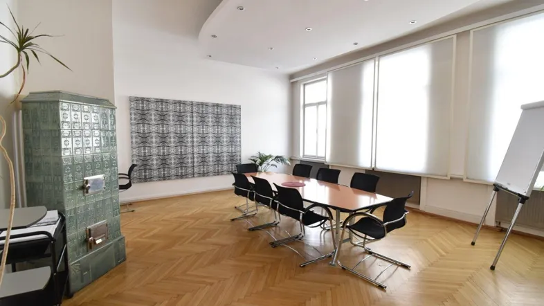 Geräumiger Büroraum mit Fischgrätparkett, großem Konferenztisch und traditionellem Kachelofen.