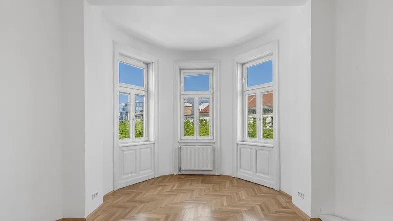 Heller Raum mit Erkerfenstern und Fischgrätparkett, ideal für Wohn- oder Arbeitsbereich.