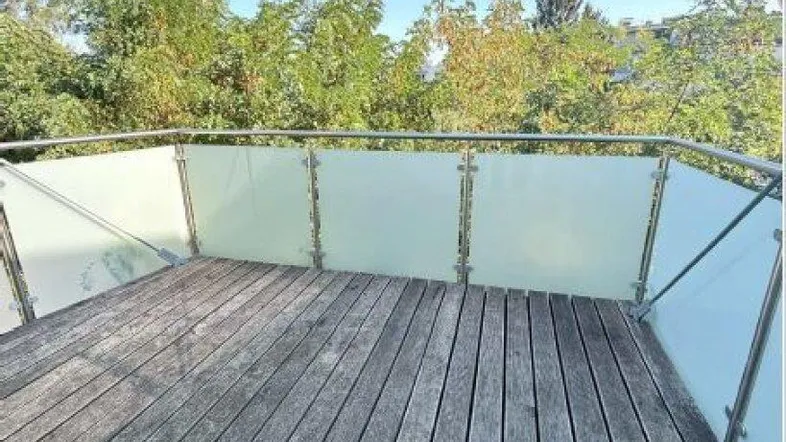 Geräumiger Balkon mit Holzdielenboden und Milchglasbrüstung, umgeben von grünen Bäumen.