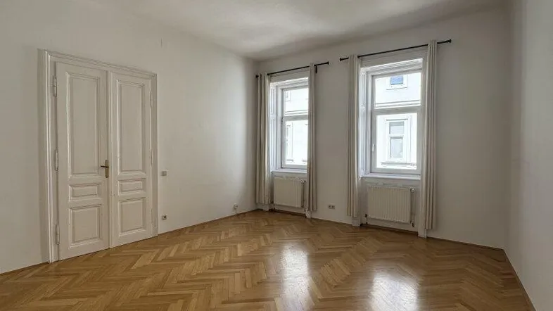Geräumiges Wohnzimmer mit hellem Fischgrätparkett, weißen Wänden und zwei großen Fenstern.