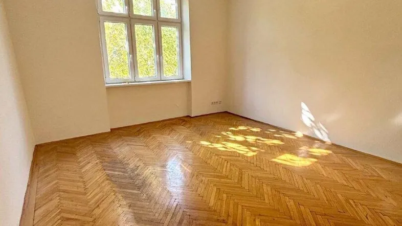 Geräumiges Zimmer mit hellem Fischgrätparkett und einem großen Fenster, das viel Tageslicht hereinlässt.