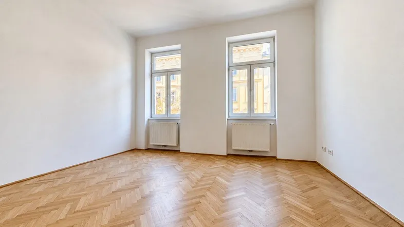 Helles Zimmer mit Fischgrätparkett und zwei großen Fenstern, ideal als Wohn- oder Schlafzimmer.