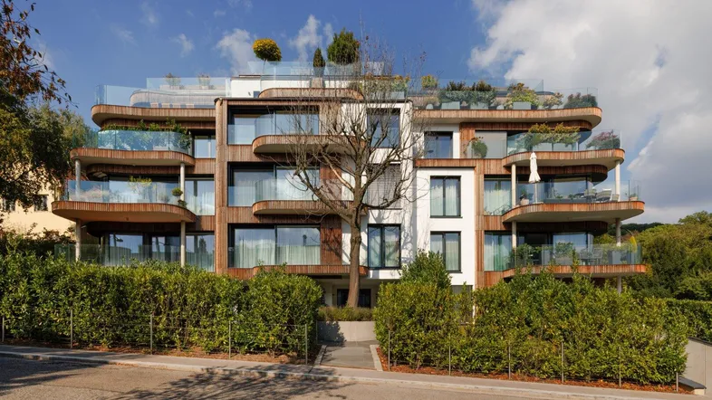 Moderne Wohnhausfassade mit Holzverkleidung, Glasbalkonen und gepflegten Hecken im Vordergrund.