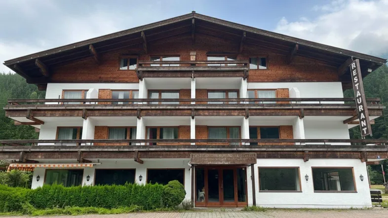 Traditionelles Gebäude mit Holzfassade und mehreren Balkonen, umgeben von grüner Landschaft und Bergen.