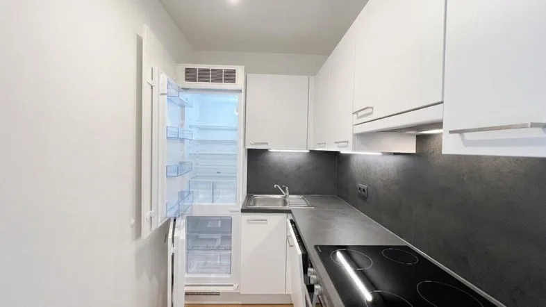 Moderne, schmale Küche mit weißen Schränken, dunkler Arbeitsplatte und integriertem Kühlschrank.