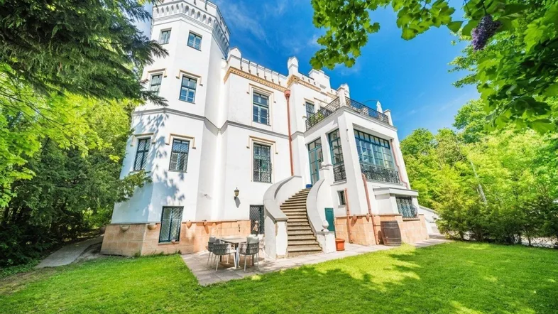 Majestätisches weißes Schlossgebäude mit Turm, umgeben von üppigem Grün und einer charmanten Außenterrasse.