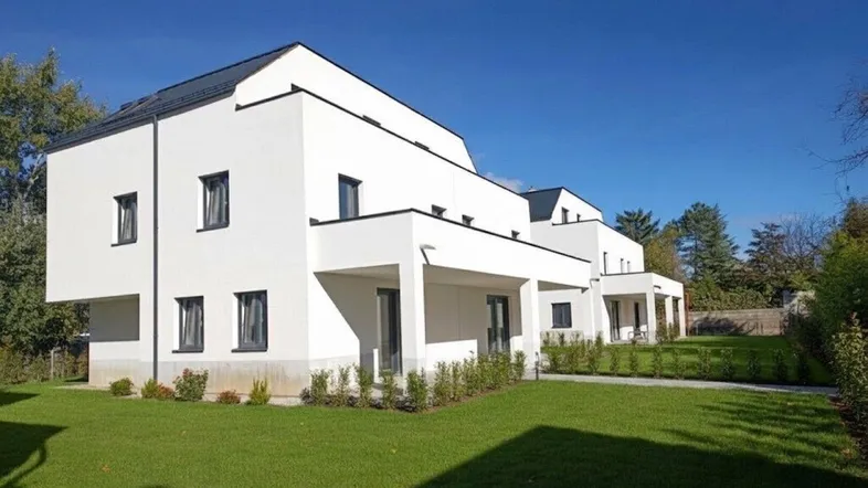 Moderne weiße Fassade eines Neubaus mit klaren Linien und gepflegtem Rasenbereich unter blauem Himmel.