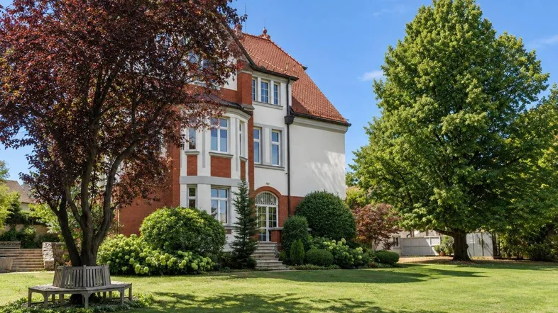 Historische Villa mit rotem Ziegeldach, gepflegtem Rasen und schattenspendenden Bäumen an einem sonnigen Tag.