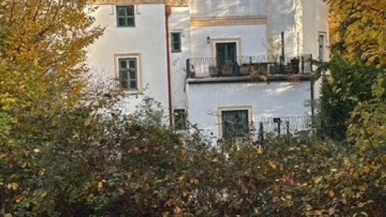 Historisches Gebäude mit Turm und Zinnen, umgeben von herbstlicher Vegetation unter blauem Himmel.