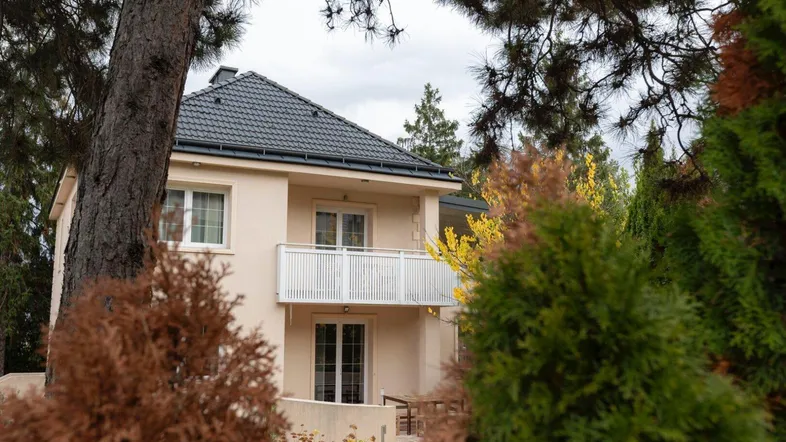 Beige Hausfassade mit Balkon und dunklem Dach, umgeben von Bäumen und Grün.