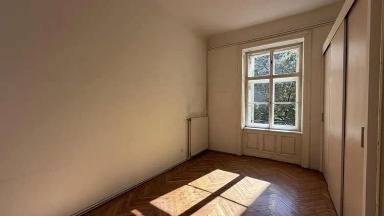 Helles Zimmer mit Fischgrätparkett und großem Fenster, das viel Sonnenlicht hereinlässt.