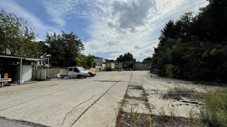 Weitläufiger Außenbereich mit gepflasterter Fläche, umgeben von Bäumen und abgestellten Fahrzeugen unter bewölktem Himmel.