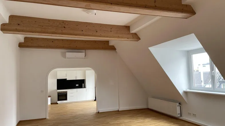 Heller Wohnbereich im Dachgeschoss mit sichtbaren Holzbalken, Parkettboden und Zugang zur Küche.