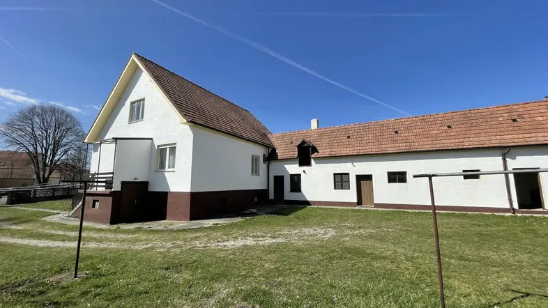 Ländliches Anwesen mit weiß getünchtem Haus, braunem Sockel und großem Rasenbereich unter blauem Himmel.