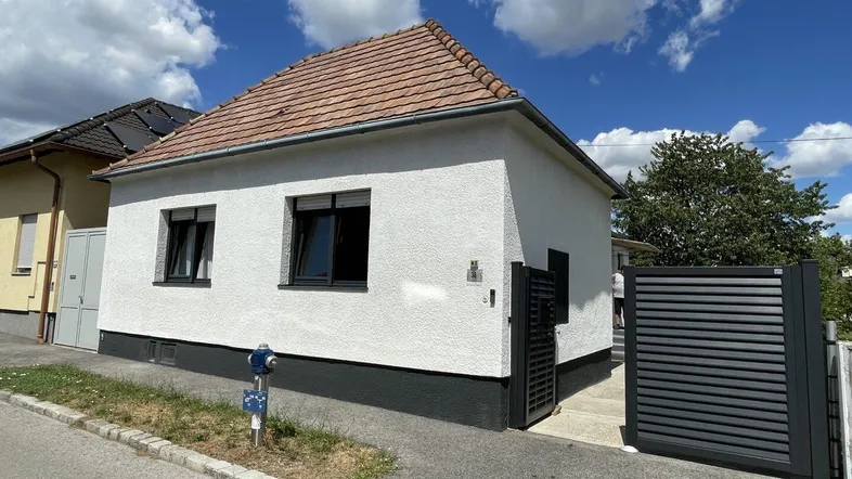 Weißes Einfamilienhaus mit Satteldach und dunklen Fenstern unter blauem Himmel.