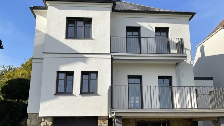 Moderne weiße Fassade eines Hauses mit dunklen Fenstern und Balkonen, teilweise mit Stein verkleidet.