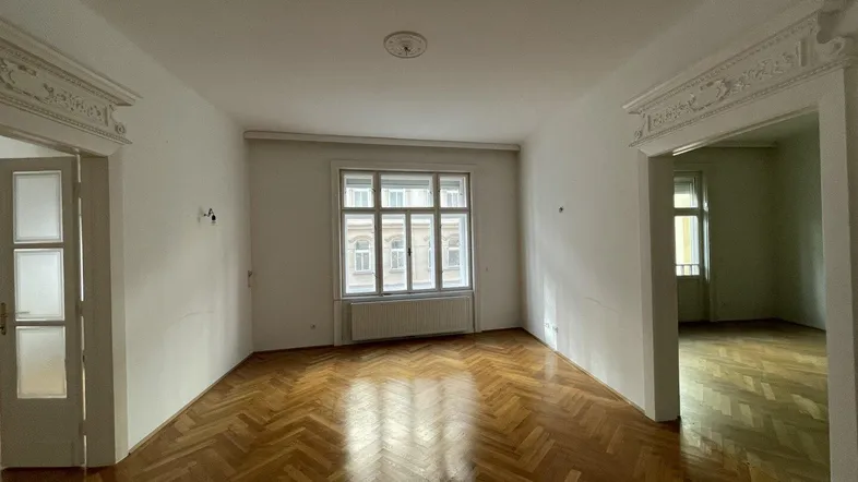 Helles Wohnzimmer mit Fischgrätparkett, hohen Decken und Stuckelementen an den Türrahmen.