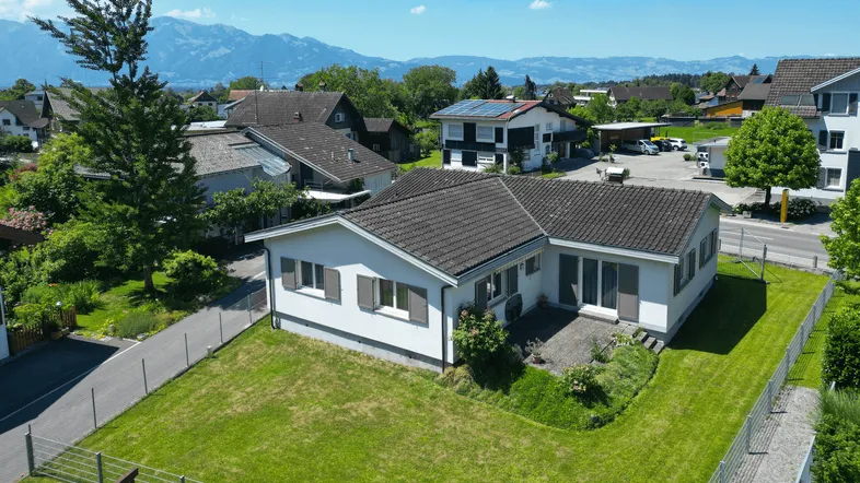 Luftaufnahme des Einfamilienhauses mit Garten und umliegender Wohngegend vor Bergkulisse.