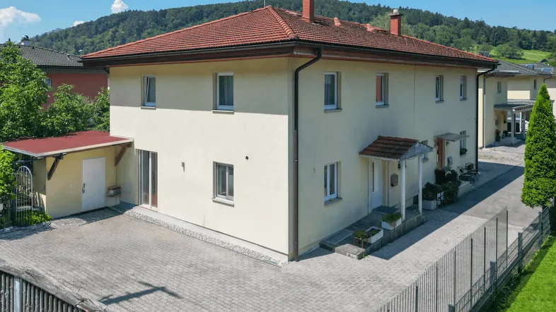 Mehrfamilienhaus mit heller Fassade, rotem Ziegeldach und gepflastertem Vorplatz vor bewaldetem Hügel.