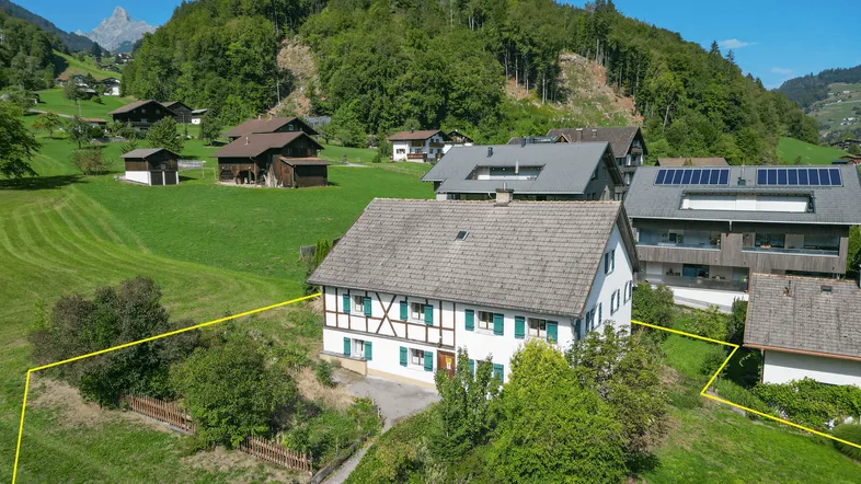 Luftaufnahme eines traditionellen Hauses mit grünen Wiesen und Bergen im Hintergrund, Grundstücksgrenzen markiert.