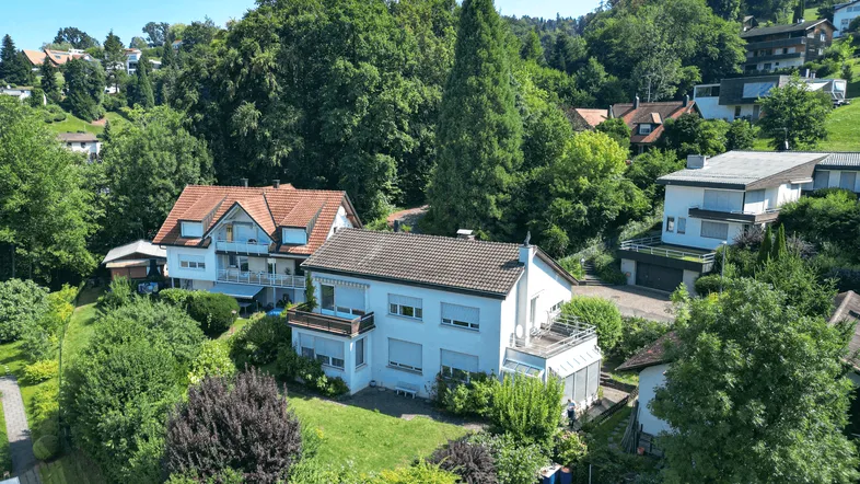 Luftaufnahme von mehreren Wohnhäusern in einer grünen, hügeligen Umgebung mit viel Baumbestand.
