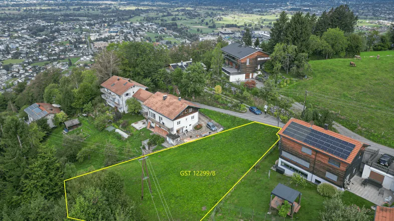 Luftaufnahme eines Grundstücks mit gelber Markierung, umgeben von Häusern und mit Fernblick auf Stadt und See.