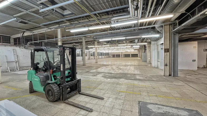 Geräumige Lagerhalle mit hohen Decken, Industrieboden und sichtbaren Lüftungsrohren, inklusive Gabelstapler.