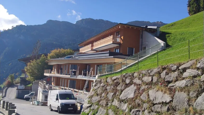 Neubauprojekt in Hanglage mit moderner Architektur und Holzfassade, umgeben von grünen Bergen.