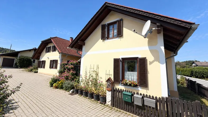 Traditionelles Haus mit gelber Fassade, braunen Fensterläden und gepflasterter Auffahrt unter blauem Himmel.