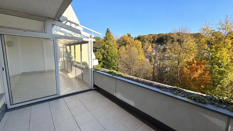 Heller Balkon mit Fliesenboden und weitem Blick ins Grüne und auf bewaldete Hügel.