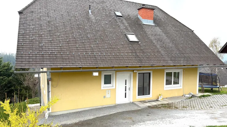 Gelbes Einfamilienhaus mit dunklem Schindeldach, umgeben von Grün und einem kleinen Gartenbereich.