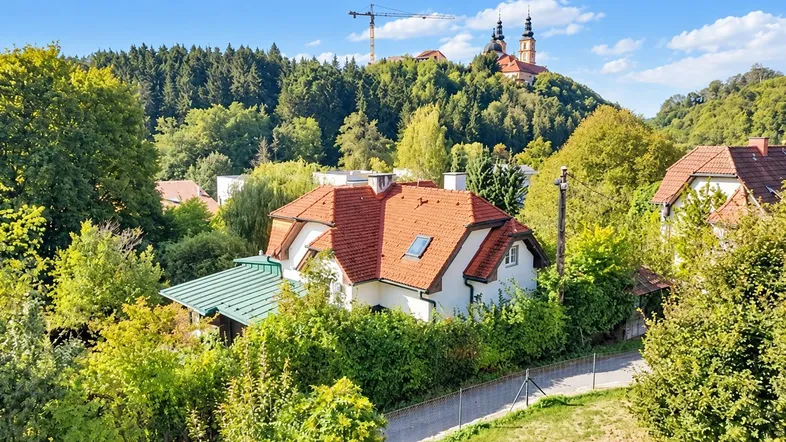 Malerisches Haus mit rotem Ziegeldach, umgeben von üppigem Grün und Hügeln.